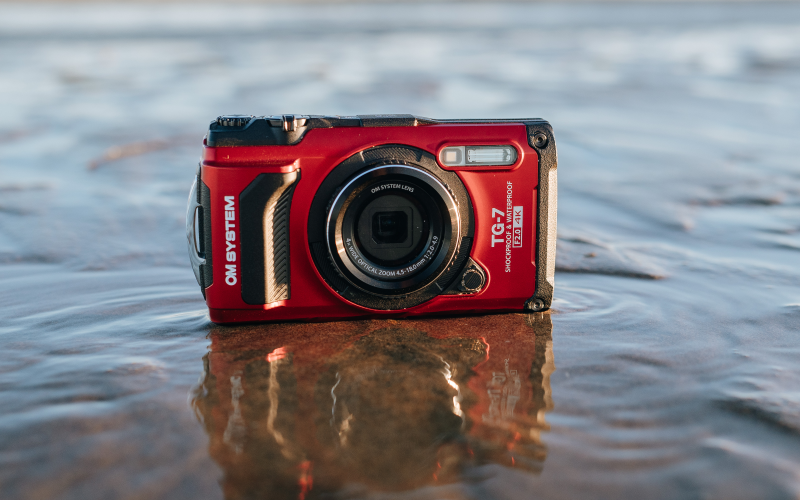 Billede af OM System TG-7 i r&oslash;d st&aring;ende i vandkanten p&aring; en strand